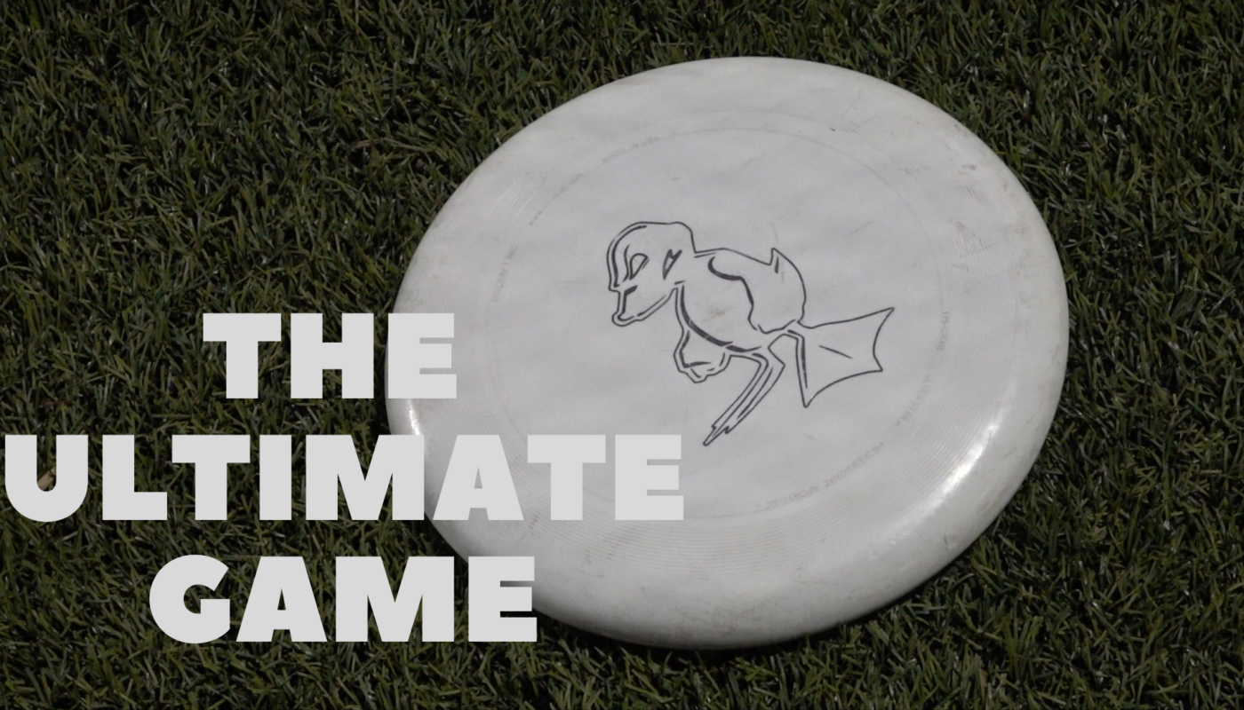 A frisbee with a duck on it lies on a turf field. The text above it reads "The Ultimate Game"