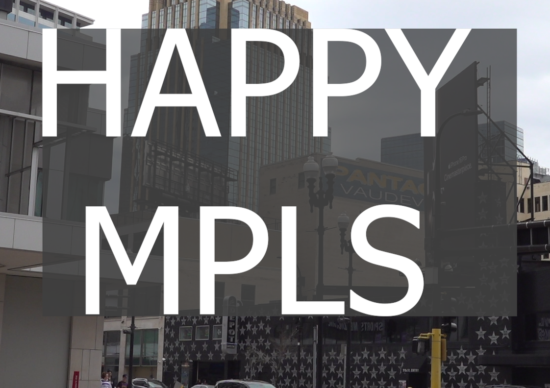 A cityscape featuring buildings, a Mayo Clinic sign, and various advertisements, including one for the iPhone 16 Cinemaster. Overlaying the image is large white text that reads "HAPPY MPLS." The scene includes cars and pedestrians, with construction cones and barriers visible on the street, suggesting ongoing roadwork. The abbreviation "MPLS" indicates that the location is Minneapolis.