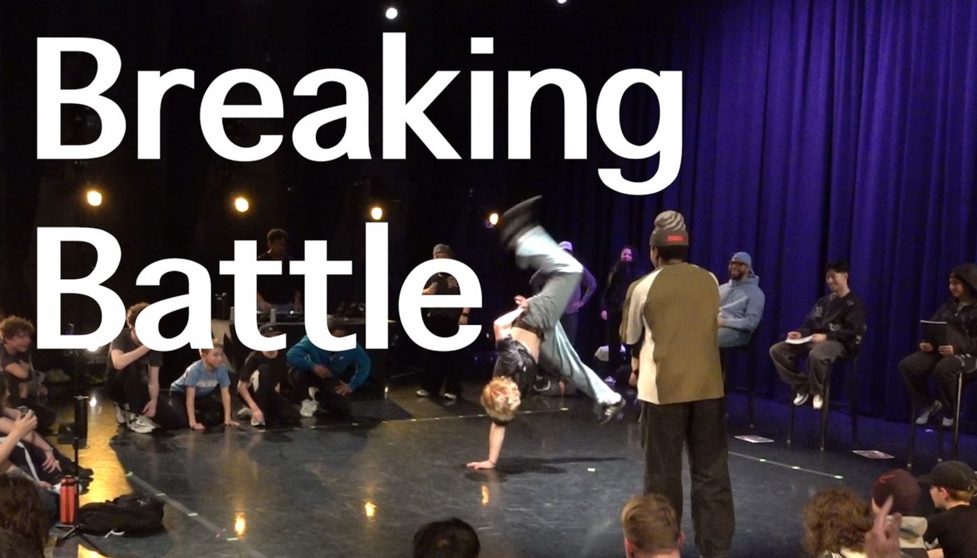 People surrounding two breakdancers as they battle each other. One is upside down on one hand and kicking his legs out. The other is watching.