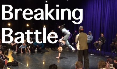 People surrounding two breakdancers as they battle each other. One is upside down on one hand and kicking his legs out. The other is watching.