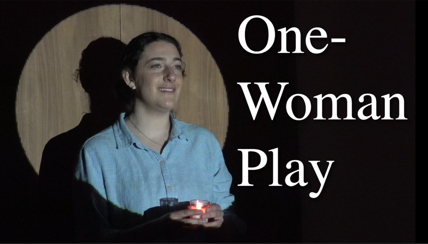 Elisheva stands in front of a spotlight casting her head and upper body in light. She holds near her chest a tea light candle in her hands.